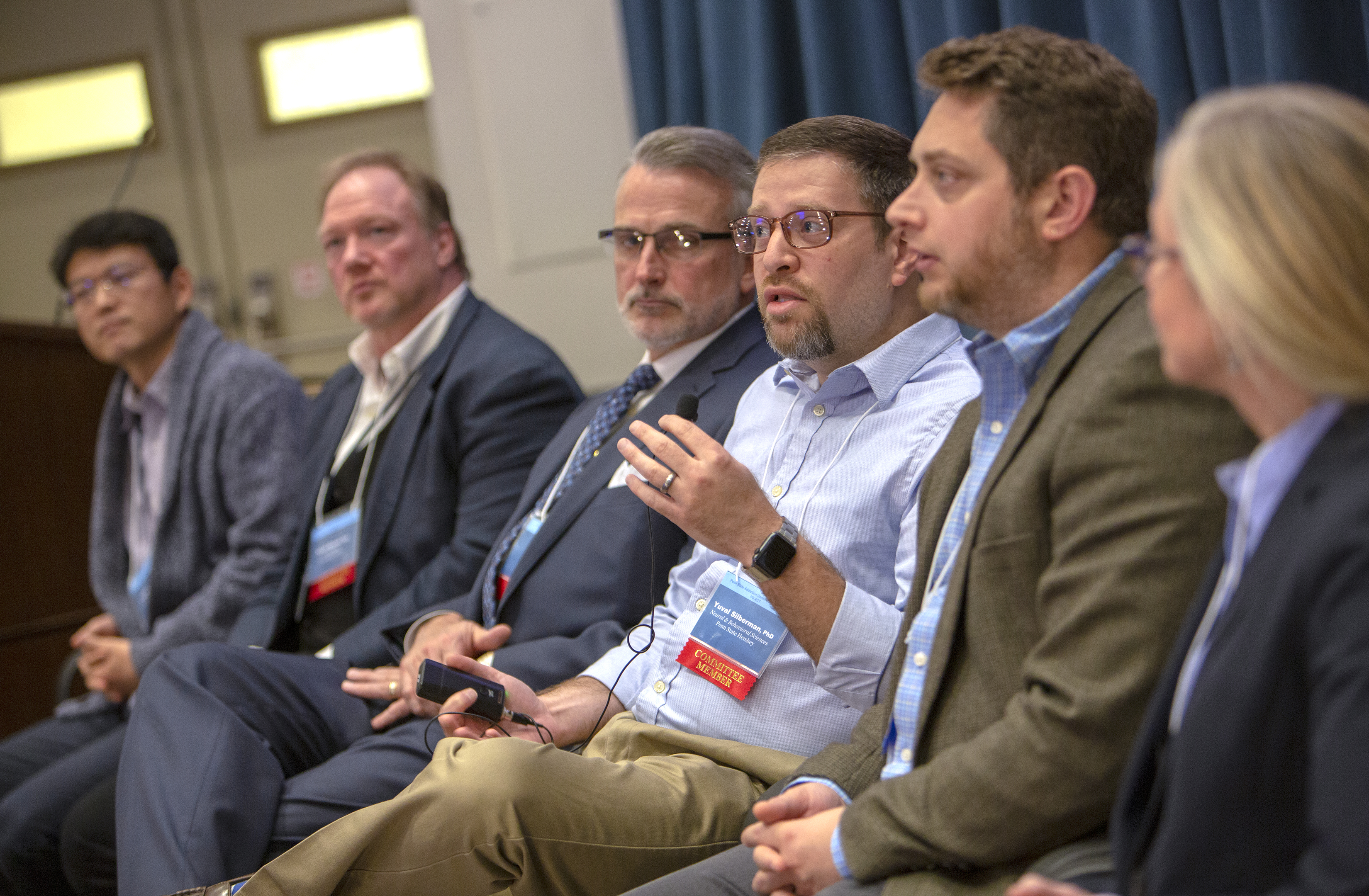 A group of six experts sit in a line during a panel discussion session. They are dressed in suits and wear committee member badges around their necks. The man third from right gestures with his hand as he speaks to the off-camera audience.