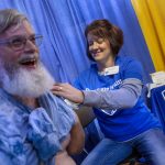 Todd Miller, a man with a long beard and glasses, smiles as a Kelly Rittle, a nurse on his right, holds up his shirt to administer a flu shot, out of the frame. Rittle is flanked by a table upon which is a rendering of the Penn State Nittany Lion in a shield.