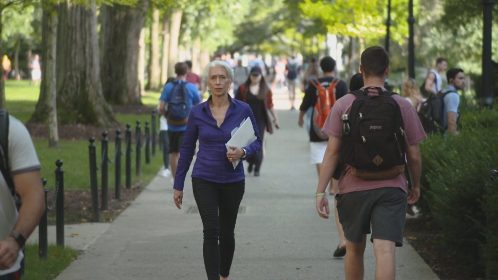 Nina Jablonski is seen walking on an outdoor pathway at Penn State University Park. She is carrying some papers in one arm, walking toward the camera. Other people are seen out of focus, going the other direction.