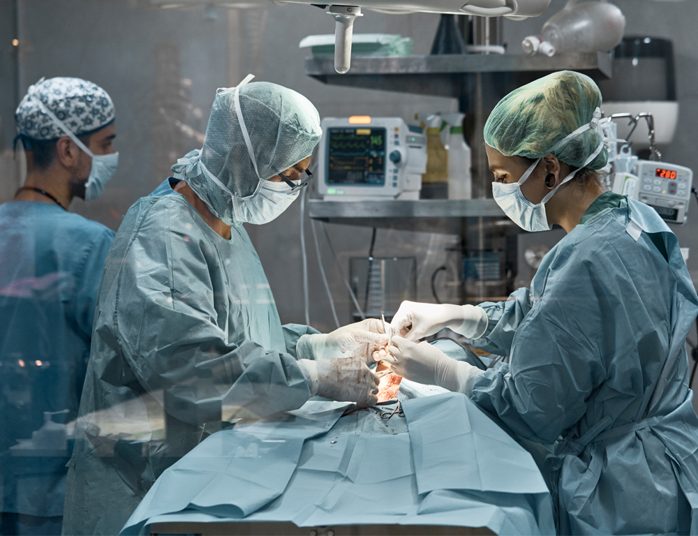 Two medical staff perform surgery on a patient. Each staff member is wearing full surgical scrubs, a mask, head covering and gloves. A large light is overhead, and various medical equipment and two other medical staffers are in the background.