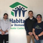 A group of faculty and students are pictured standing against a wall with the Habitat for Humanity logo on it.