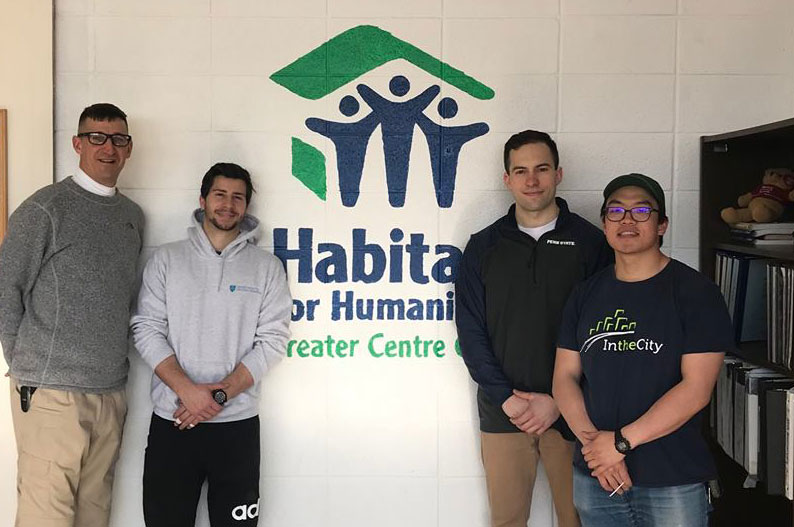 A group of faculty and students are pictured standing against a wall with the Habitat for Humanity logo on it.