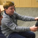A teen boy smiles and holds the wheel of the driving simulator One Simple Decision. Next to him is a man with glasses, gray hair and a beard. Behind them are a table and chairs, carpet and a wooden cabinet.