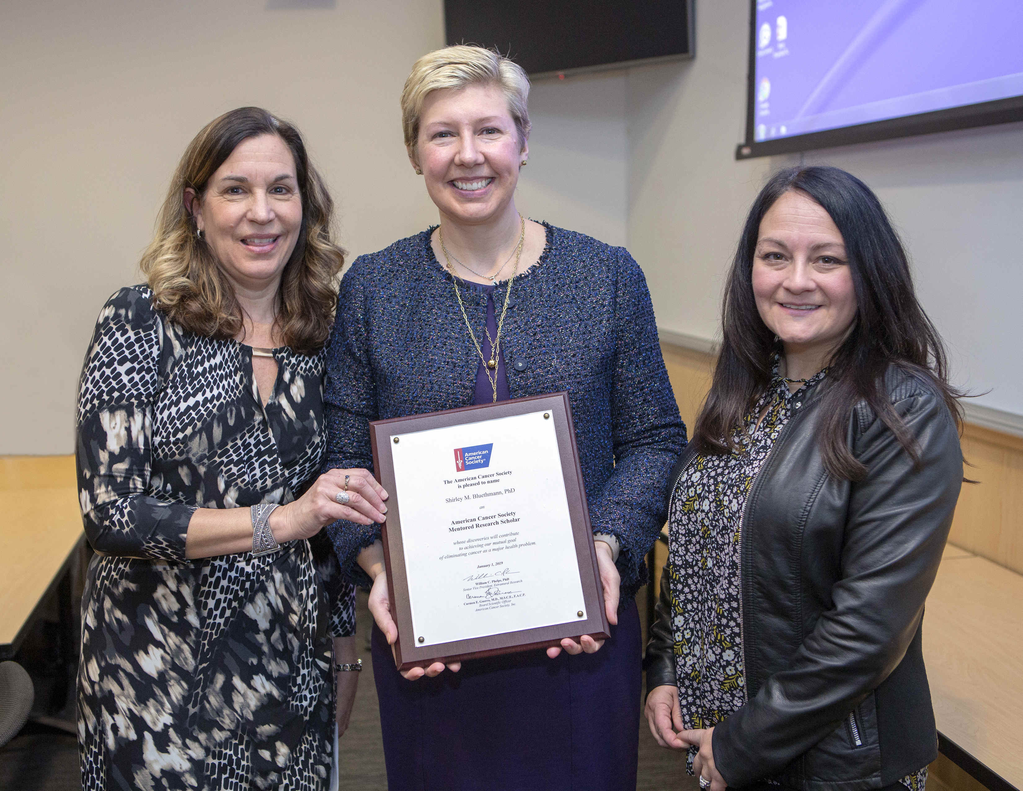 Three women in professional attire pose for a photo in a conference room. The woman in the middle holds a framed certificate bearing the American Cancer Society logo. A portion of a table and parts of two wall-mounted video monitors are in the background.