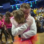 A young man and a child embrace on the floor of THON at the Bryce Jordan Center. The man, facing the camera, is smiling. Several people are in the background – some standing, some seated, slightly out of focus. A stage is also visible.