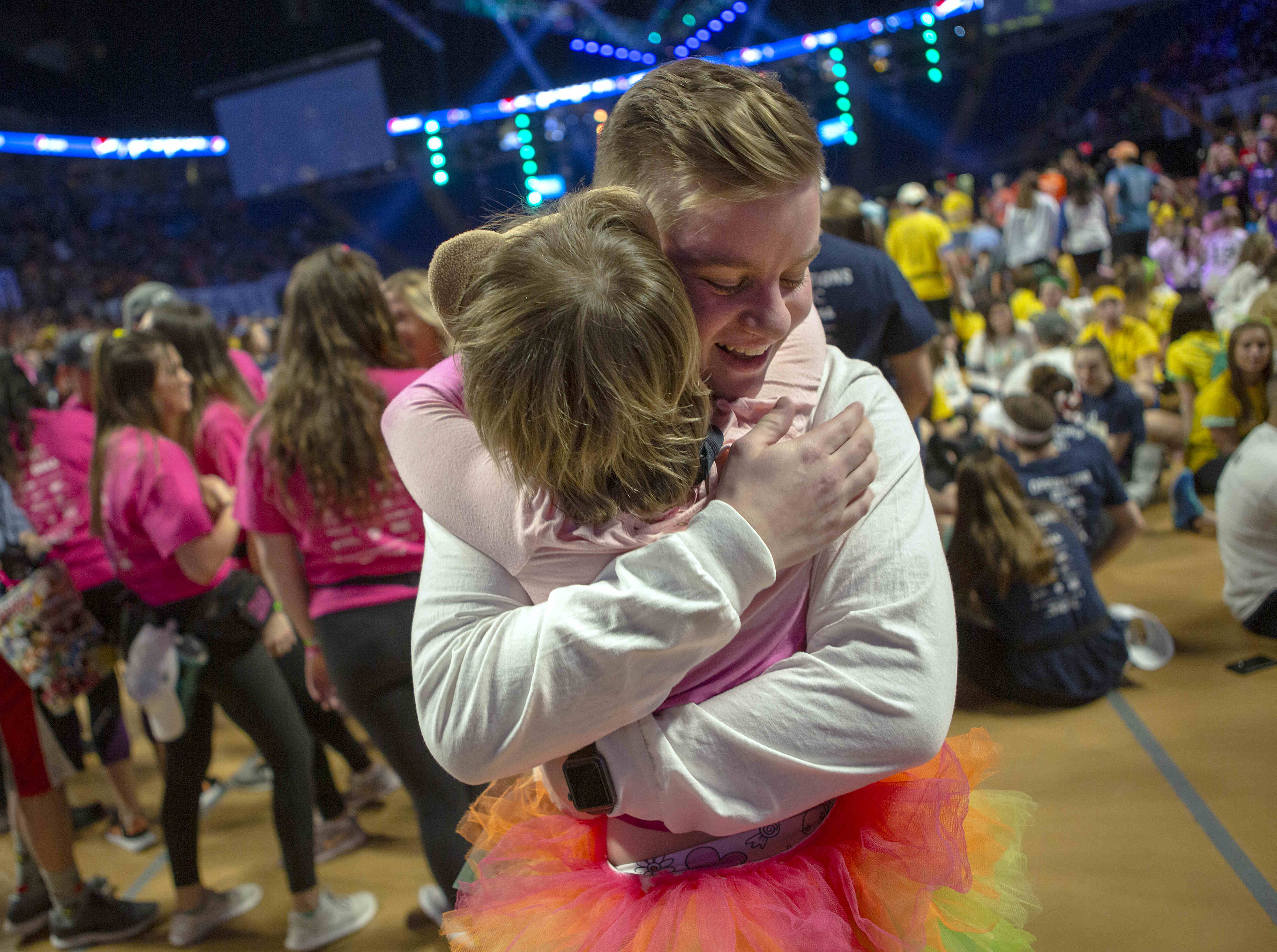 A young man and a child embrace on the floor of THON at the Bryce Jordan Center. The man, facing the camera, is smiling. Several people are in the background – some standing, some seated, slightly out of focus. A stage is also visible.