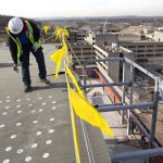 To workers in hard hats complete work on the top floor of a building. To their right, scaffolding partially obscures a look across rooftops and a skyline.