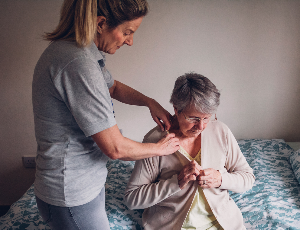 A woman sits on the edge of a bed, buttoning a sweater. Another woman stands at her side, helping her put the sweater on.