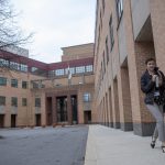 A young woman walks on a sidewalk outside 100 Crystal A Drive, Penn State Health’s new corporate headquarters. The brick building is designed in a U-shape. A flag is over the entrance. A bare tree and pile of snow are on the left. The woman is wearing a short jacket, casual pants, high heels and holding a backpack from her arm.