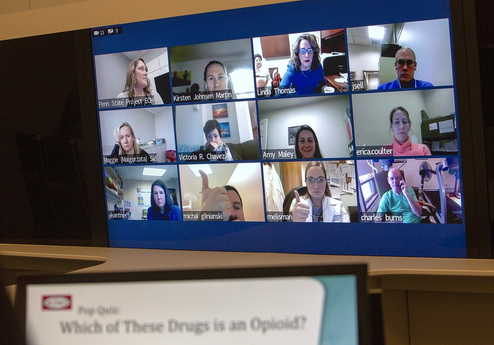 A large screen contains 12 mini screens showing physicians participating in Project ECHO. The participants are looking into their own web cams as they participate in the session; some are smiling and giving a thumbs up while others are serious. In the foreground, an out-of-focus screen reads “Pop Quiz: Which of these drugs is an opioid?”
