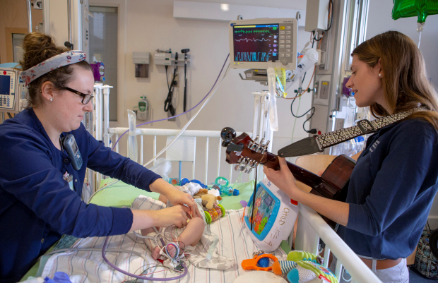 Marissa Aulenbach, right, a board-certified music therapist at Penn State Health Children's Hospital, plays a guitar while registered nurse Lauren Libhart tends to 4-month-old Caden Hoover. The baby is lying in a crib with wires and monitors attached to his body. Aulenbach is wearing a blue, long-sleeved shirt and jeans. Libhart is wearing blue scrubs, a headband and glasses. Several toys are in the crib.