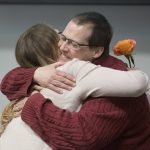 Ashley Brewer receives a hug from organ recipient Bill Griffis at the Rose Parade Donor Remembrance Ceremony. Ashley is a young woman with long hair wearing a sweater and holding a rose. Bill is smiling and has his eyes closed. He is a middle-aged man wearing glasses and a sweater.