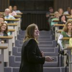 Deborah Berini, president of Penn State Health Milton S. Hershey Medical Center, smiles while speaking at an employee meeting in November. She is seen in profile as she turns right and aims a remote to advance a slide. Rows of employees sit in rows in Junker Auditorium’s raised seating. Carpeted steps are behind Deborah.