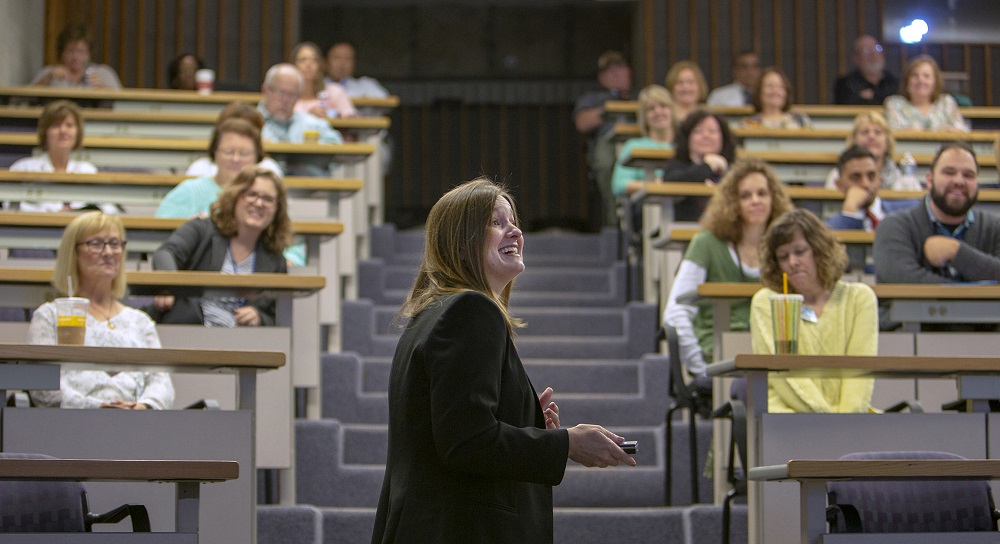Deborah Berini, president of Penn State Health Milton S. Hershey Medical Center, smiles while speaking at an employee meeting in November. She is seen in profile as she turns right and aims a remote to advance a slide. Rows of employees sit in rows in Junker Auditorium’s raised seating. Carpeted steps are behind Deborah.