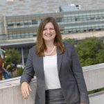 Deborah Berini, president of Penn State Health Milton S. Hershey Medical Center, leans on a concrete wall overlooking Penn State Health Children's Hospital. She is wearing a suit, top and a pearl necklace. She has straight, shoulder-length hair and is smiling. A colorful metal sculpture is in front of the Children’s Hospital.