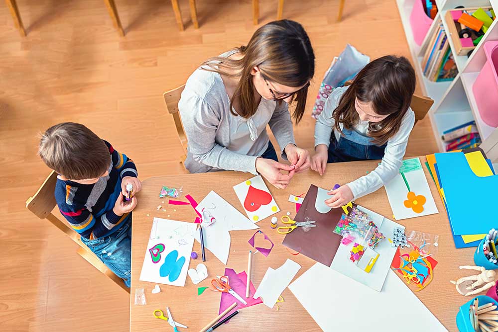 A woman sits between two children at a table, holding something between her fingers. The table is covered with construction paper, scissors, pens and other craft tools.