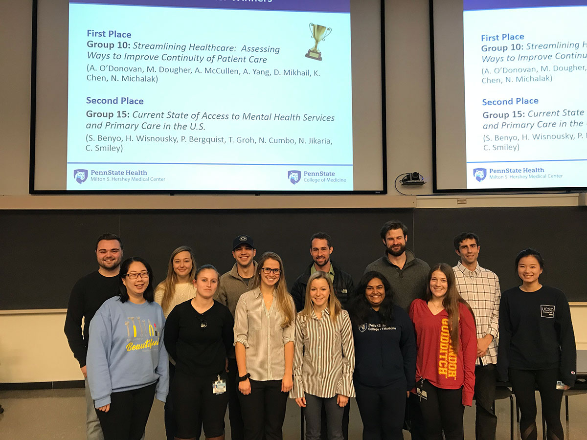 A large group of students is pictured standing in a lecture room, in front of projector screens listing various poster topics.