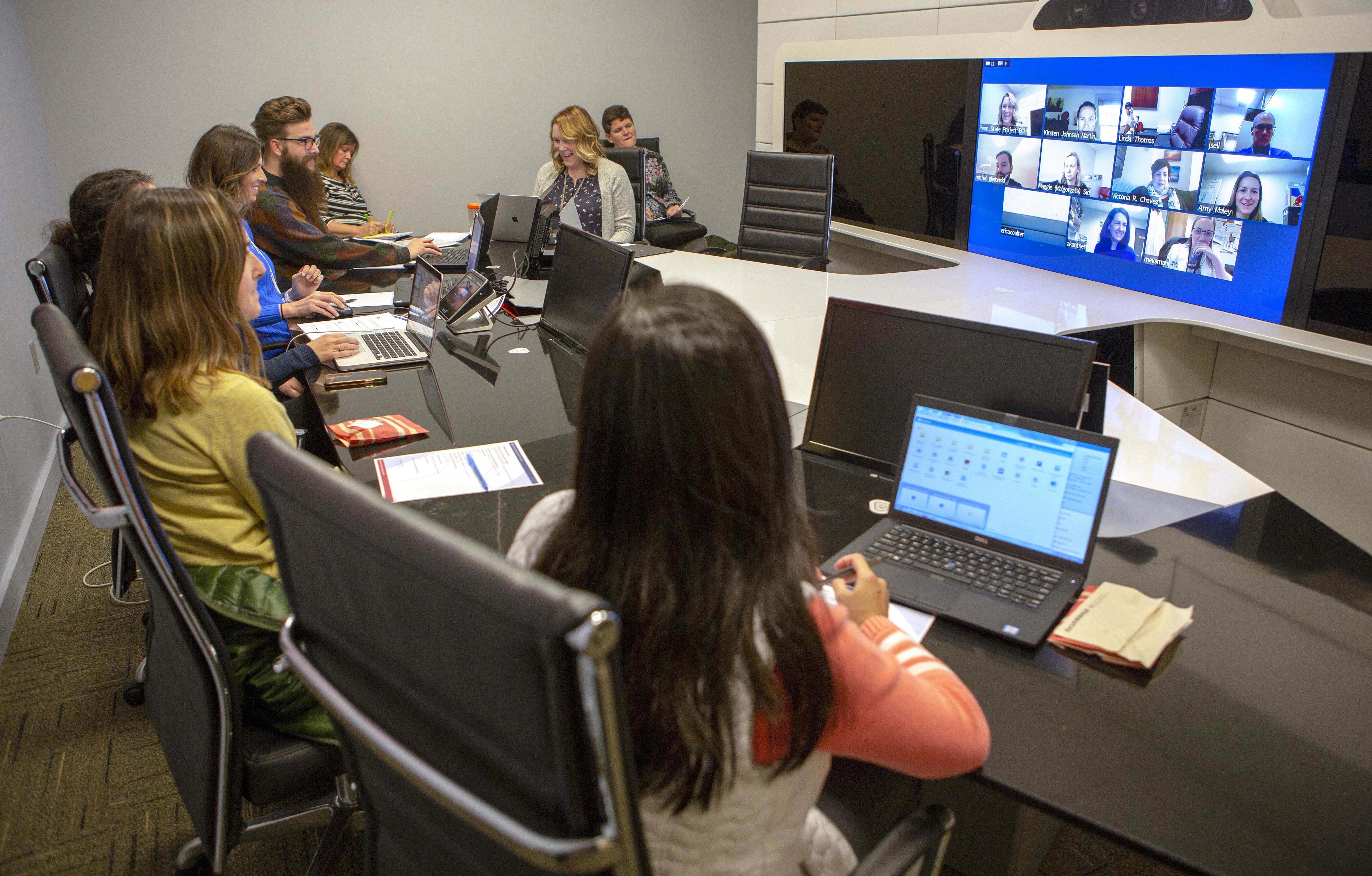 A long office table has eight people seated on one side and at its head. They have laptops open in front of them and also face a screen projected onto a panel on the opposite wall. The projection shows a series of mini screens of other participants in the Project ECHO teleconference. People are smiling.