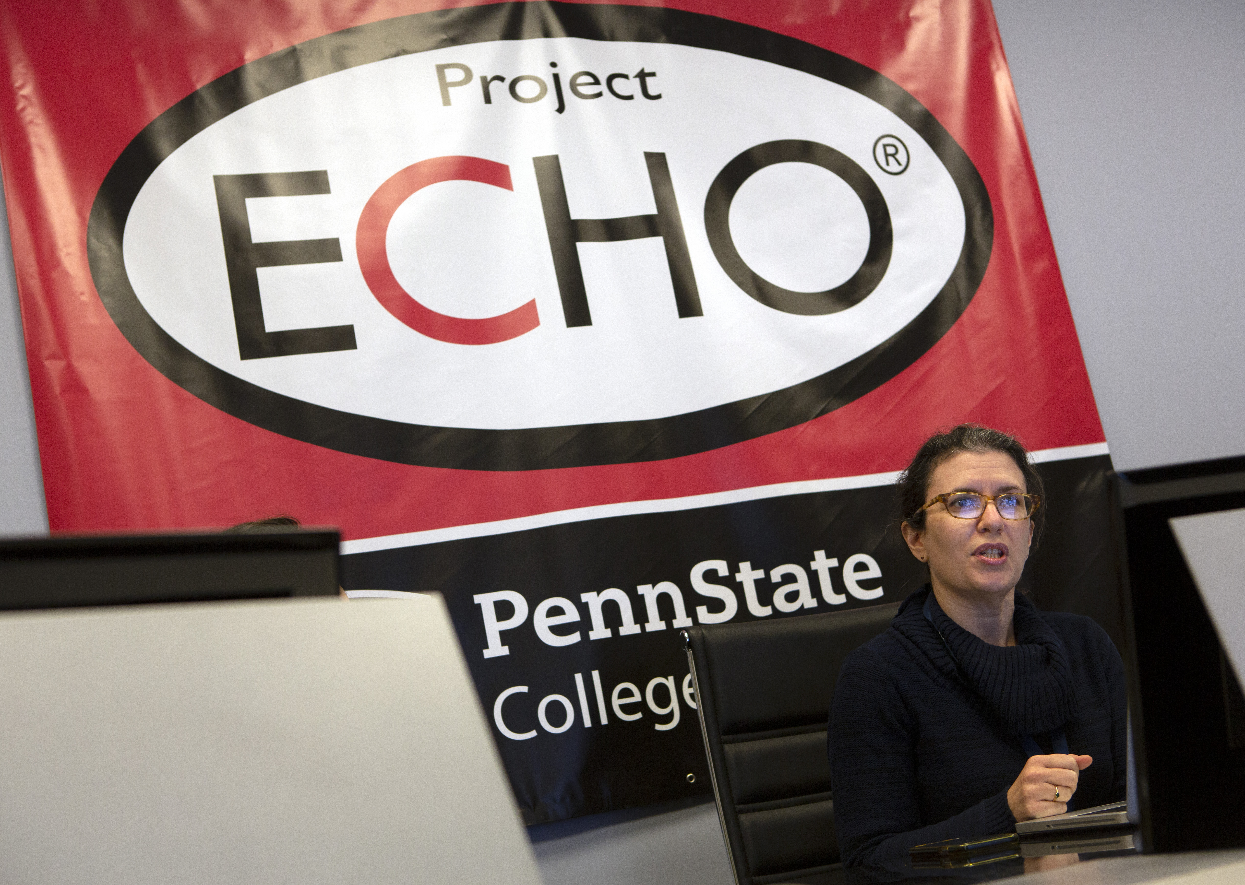 A woman with a black turtleneck sweater and glasses is seated at a table with a laptop open in front of her. She is speaking, facing a lit-up screen that is reflected in her glasses. Behind her is a large red, black and white sign reading “Project ECHO” with portions of “Penn State College of Medicine” visible. Another person is seated in the lower left corner but is mostly hidden by another computer screen.