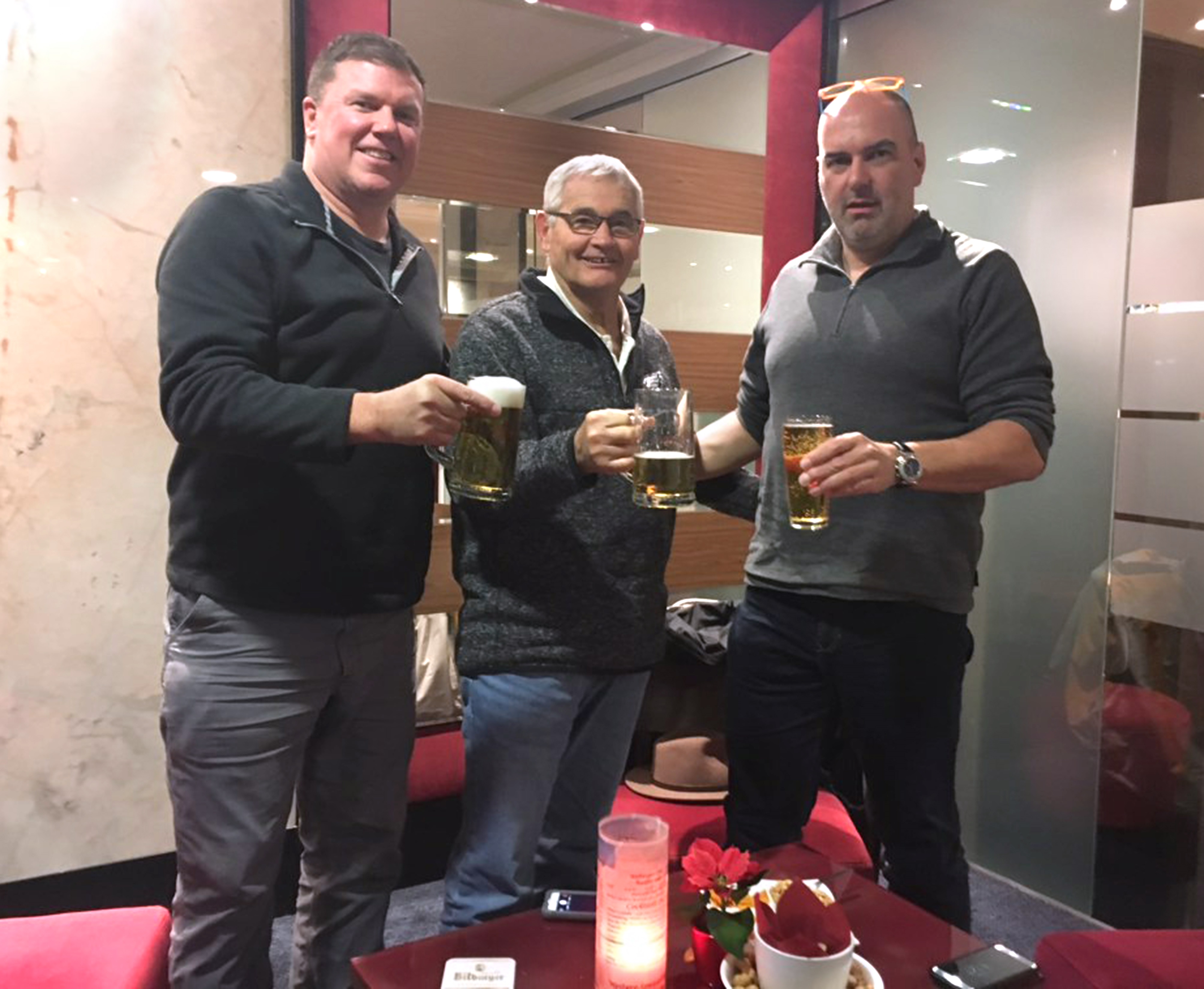 Jim Miller, center, toasts his donor, Stefan Eichert, right, with John Moore, left, the businessman who invited Miller to accompany him on a trip to Germany. All three men are wearing casual shirts and jeans and holding mugs of beer. A candle is on the table in front of them. Behind them is a marbled wall and a wall decorated with slats of wood.