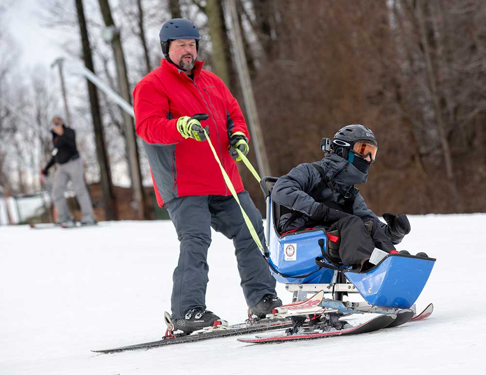 A man in a parka on a pair of skis holds onto straps behind a man in goggles and winter wear in a plastic seat outfitted with skis. Both men are sliding on snow with bare trees in the background.