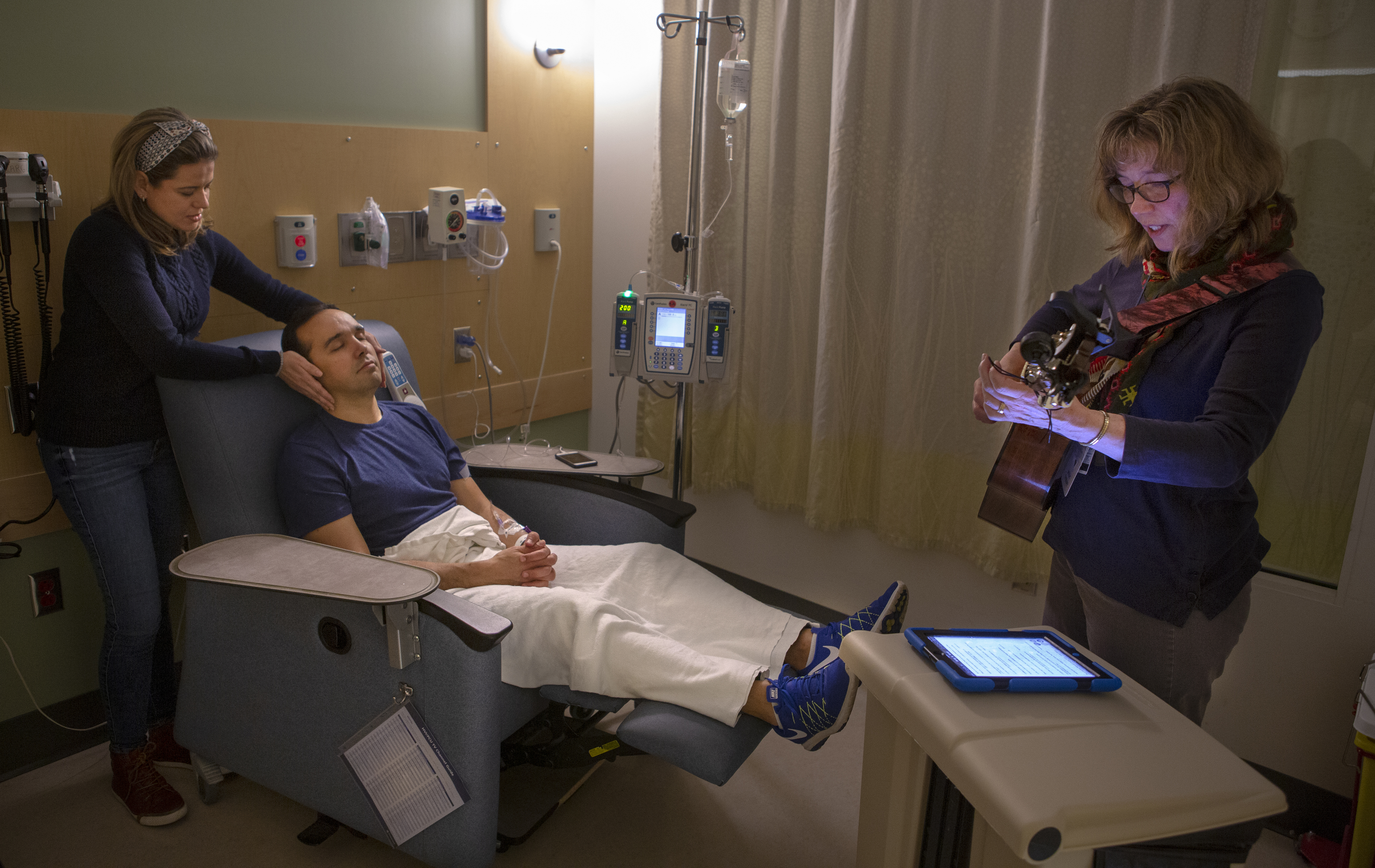 Jan Stouffer, a board-certified music therapist at Penn State Health Milton S. Hershey Medical Center, plays the guitar for Vladimire de Abreu and his wife, Samia, during an infusion session at Penn State Cancer Institute. Stouffer is wearing a blue, long-sleeved shirt and khaki pants. Samia has her hands around her husband’s face as he lies in a chair with his eyes closed. She is wearing a blue shirt and jeans. Vladimire is lying with his feet up in a recliner and has an IV in his arm. His is wearing a blue T-shirt and white pants.