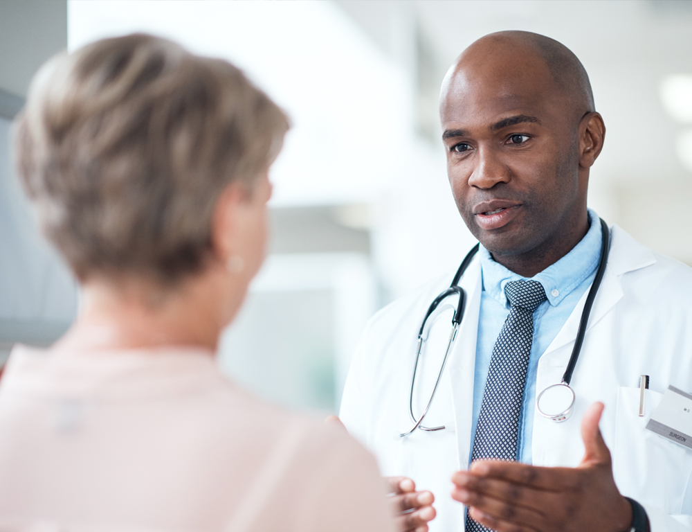 A male physician, wearing a stethoscope around his neck and a white coat, gestures with his hands as he speaks to a woman facing him. He faces the camera; she is facing away from it.