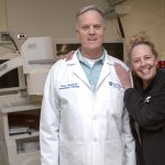 Bobbie Mann of Lebanon, Pa., smiles with her arm around Dr. Thomas McGarrity, a gastroenterologist at Penn State Health Milton S. Hershey Medical Center. She has blonde hair and is wearing a sweatshirt. Dr. McGarrity is wearing a white lab coat with the Medical Center logo on the right and his name and “Gastroenterology & Hepatology” on the left. They are standing in front of a colonoscope and three monitors.