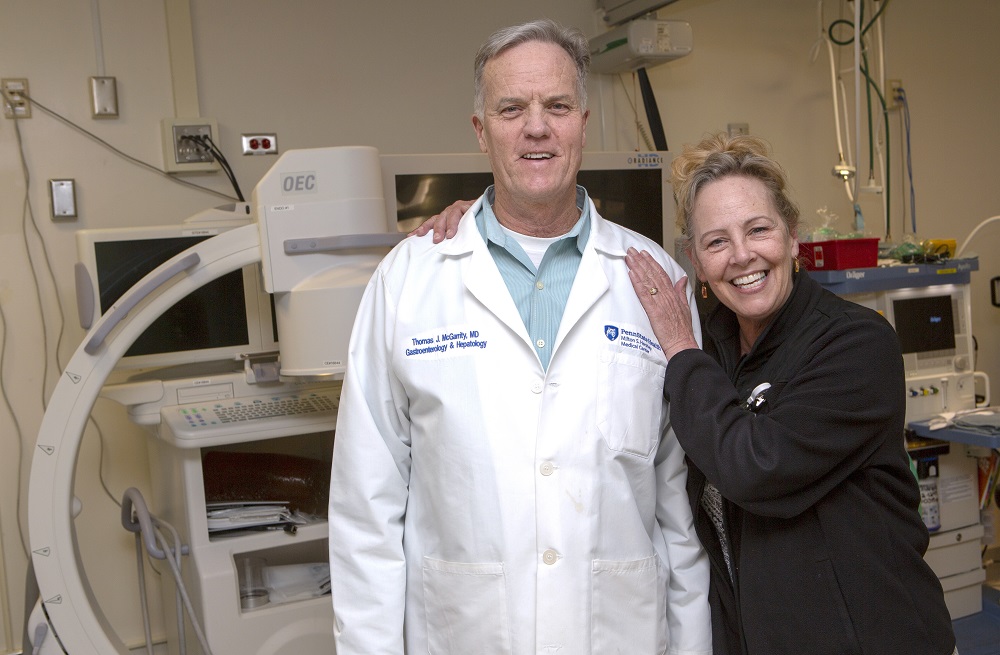 Bobbie Mann of Lebanon, Pa., smiles with her arm around Dr. Thomas McGarrity, a gastroenterologist at Penn State Health Milton S. Hershey Medical Center. She has blonde hair and is wearing a sweatshirt. Dr. McGarrity is wearing a white lab coat with the Medical Center logo on the right and his name and “Gastroenterology & Hepatology” on the left. They are standing in front of a colonoscope and three monitors.