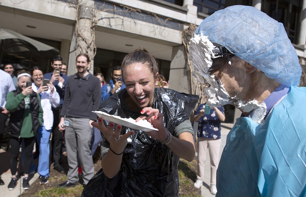 Dr. Michael Katzman, right, stands with whipped cream on his face and sunglasses during the Pi Day fundraiser. He is wearing a paper smock and hair net. First-year medical student Madelaine Fritsche laughs as she holds a plate filled with whipped cream. Behind them a large group of onlookers laughs and takes photos with their cell phones. They are standing outdoors in a courtyard.