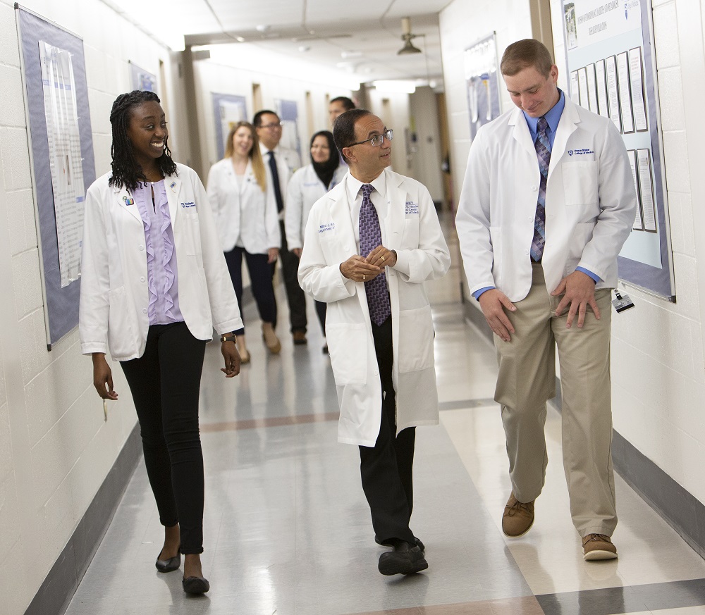 Two men and a woman in white lab coats walk through a tile-floored hallway. Another group of four people are in the background behind them.