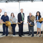 Seven people in formal attire hold gold shovels and toss dirt in the air. A large rendering of Penn State Health Hampden Medical Center is in the near background.