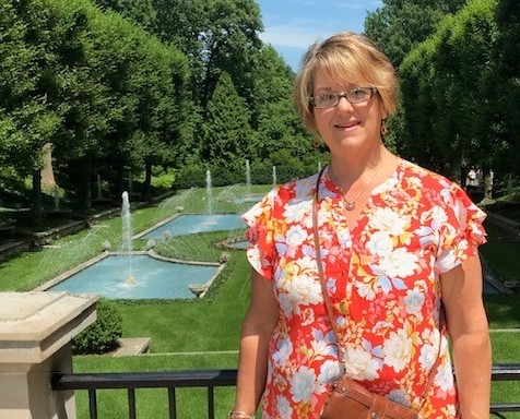 A woman wearing an orange blouse with white flower pattern stands in front of a railing with a green lawn and fountains in the background.