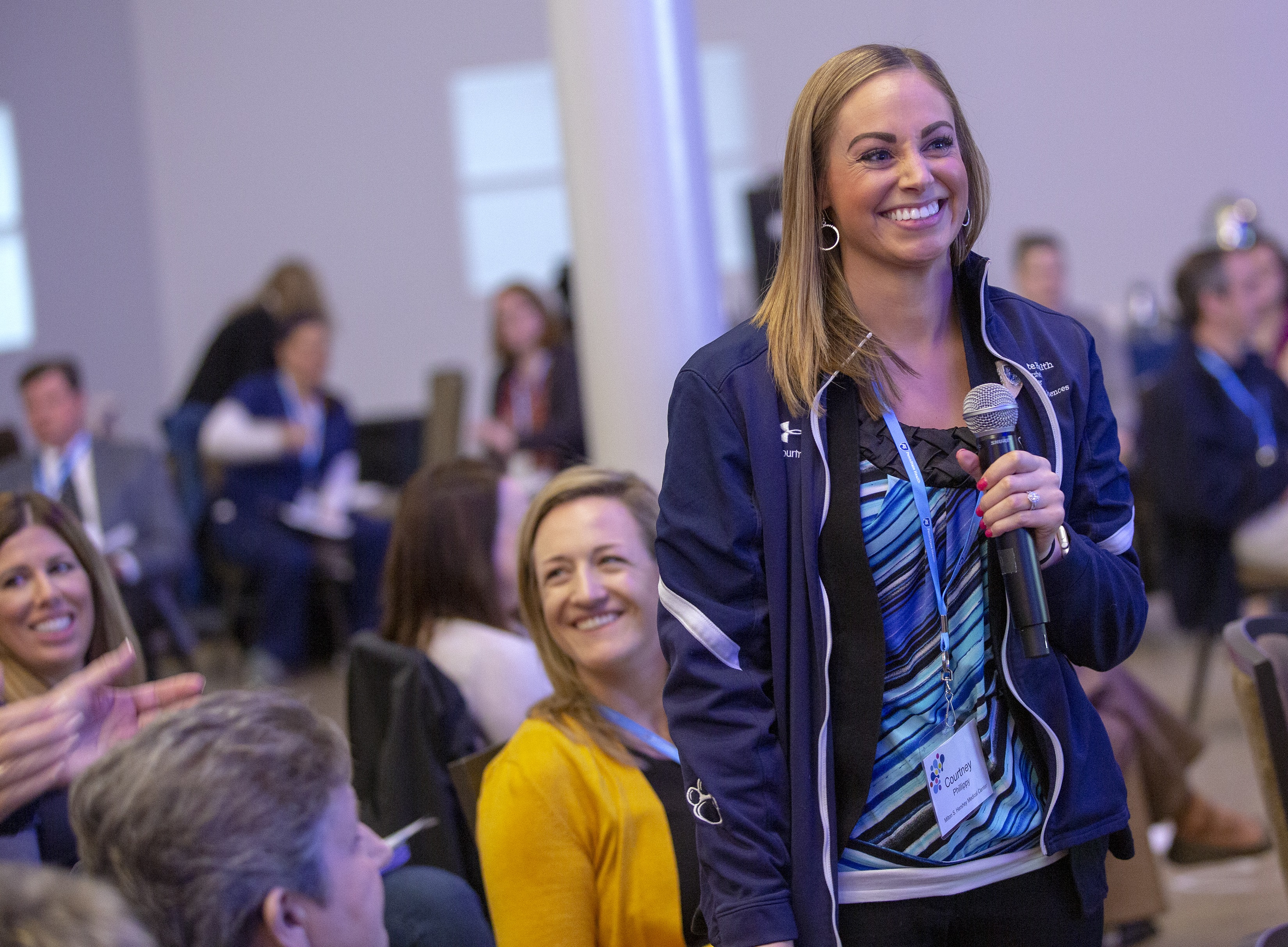 Courtney Phillippy, a woman with long, blonde hair, stands with a microphone in her hand and smiles during a small group session at the 2019 Leadership Conference: This is Penn State Health. She is wearing a Penn State jacket and a striped top. Behind her other employees smile or look to the side. A large steel column is behind her.