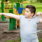 A young boy stands with his right arm extended outward and his left arm curled upward. Playground equipment is in the background.