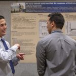 Two men speak to one another in front of a poster with the words “All trans-retinoic acid modifies the response of A549 alveolar epithelial cells to hyperoxia. The man wears a tie and gestures. The man on the right is facing the poster with his head turned toward the gesturing man.