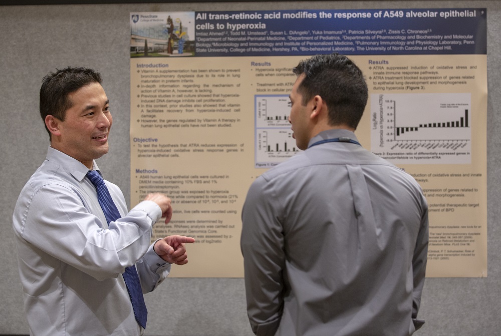 Two men speak to one another in front of a poster with the words “All trans-retinoic acid modifies the response of A549 alveolar epithelial cells to hyperoxia. The man wears a tie and gestures. The man on the right is facing the poster with his head turned toward the gesturing man.