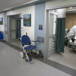 A physician and two other care providers treat a patient who is lying in a bed in Hershey Medical Center’s new Radiology Outpatient Care Unit. A long hallway with a tiled floor shows five rooms. The care providers and patient are in the room on the right, which has a curtain and a door. A wheelchair is outside the door. Paintings are on the walls between each room. Medical waste containers are outside each room.
