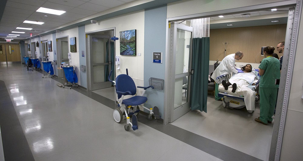 A physician and two other care providers treat a patient who is lying in a bed in Hershey Medical Center’s new Radiology Outpatient Care Unit. A long hallway with a tiled floor shows five rooms. The care providers and patient are in the room on the right, which has a curtain and a door. A wheelchair is outside the door. Paintings are on the walls between each room. Medical waste containers are outside each room.