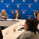 Pennsylvania Health Secretary Dr. Rachel Levine speaks in front of a screen featuring the Penn State Health logo, while seated at a conference table. Other men and women seated at the table watch her.