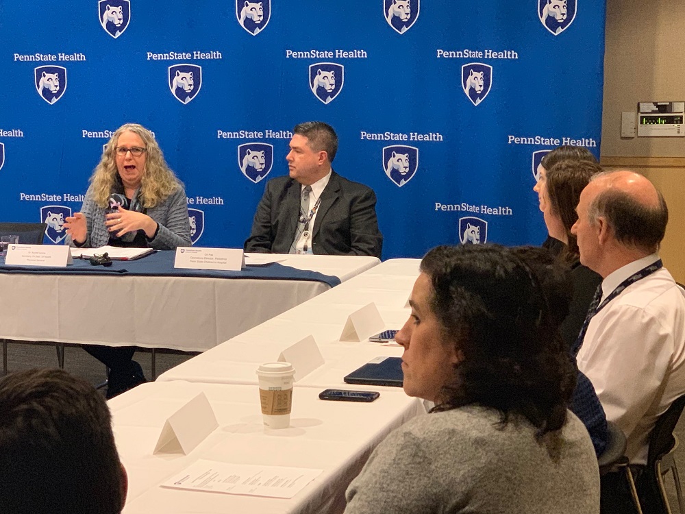 Pennsylvania Health Secretary Dr. Rachel Levine speaks in front of a screen featuring the Penn State Health logo, while seated at a conference table. Other men and women seated at the table watch her.