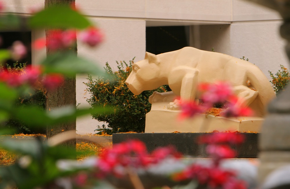 A statue of the Nittany Lion looks toward some flowering bushes. In the foreground, flowers are blooming.