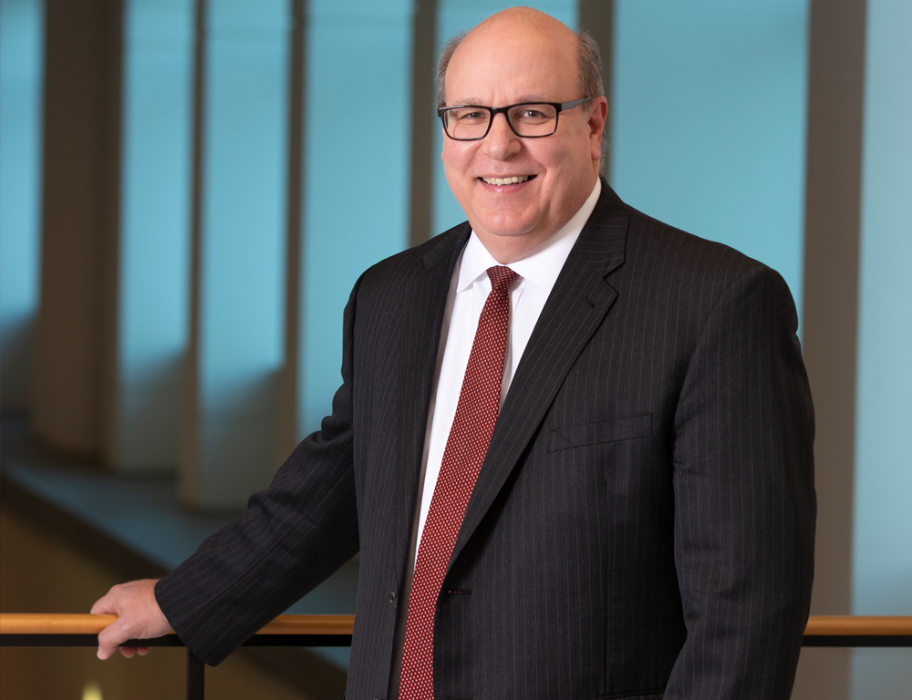 Penn State Health CEO Steve Massini is shown in a professional headshot wearing a suit, tie and glasses. He is standing on a balcony holding a railing. Behind him is a bank of windows.