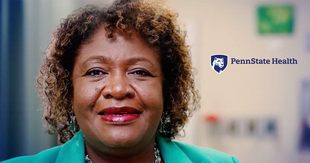 Wanda Johnson of Information Services at Penn State Health smiles at the camera. She has curly hair and is wearing a suit jacket and silver necklace. On her right is the Penn State Health logo over an out-of-focus wall.