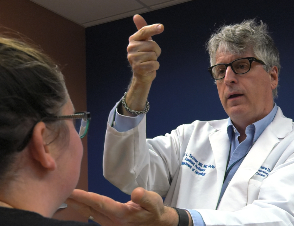 Dr. Cary Twyman, a neurologist at Penn State Health, examines a patient. He stands in front of her, his left hand on her chin, his right hand extended slightly into the air.