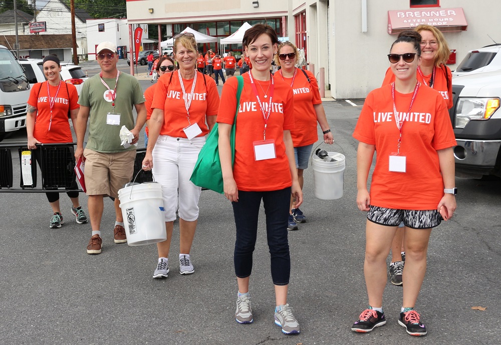 Several women and a man in shorts, T-shirts, yoga pants or clam digger pants carry buckets, stepladders and equipment as they make their way through a car-dealership parking lot. Most of the T-shirts say Live United.