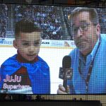 A boy in a cape appears on a video screen bracketed by candy ads. A man with a microphone appears to the left of the boy. Both are positioned on an ice rink with a hockey player partially visible behind them.