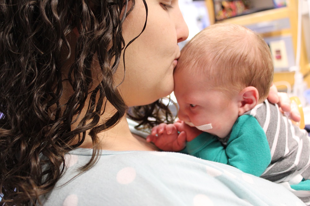 Natasha Himes kisses her newborn baby boy as she holds him on her chest. He has surgical tape on his face and his wearing a striped shirt. Natasha has long curly hair and is wearing a cotton top.