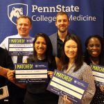 A group of seven people is seen standing in front of a Penn State College of Medicine banner. The students in the middle are holding signs that say "I Matched!" and that list residency location and specialty.
