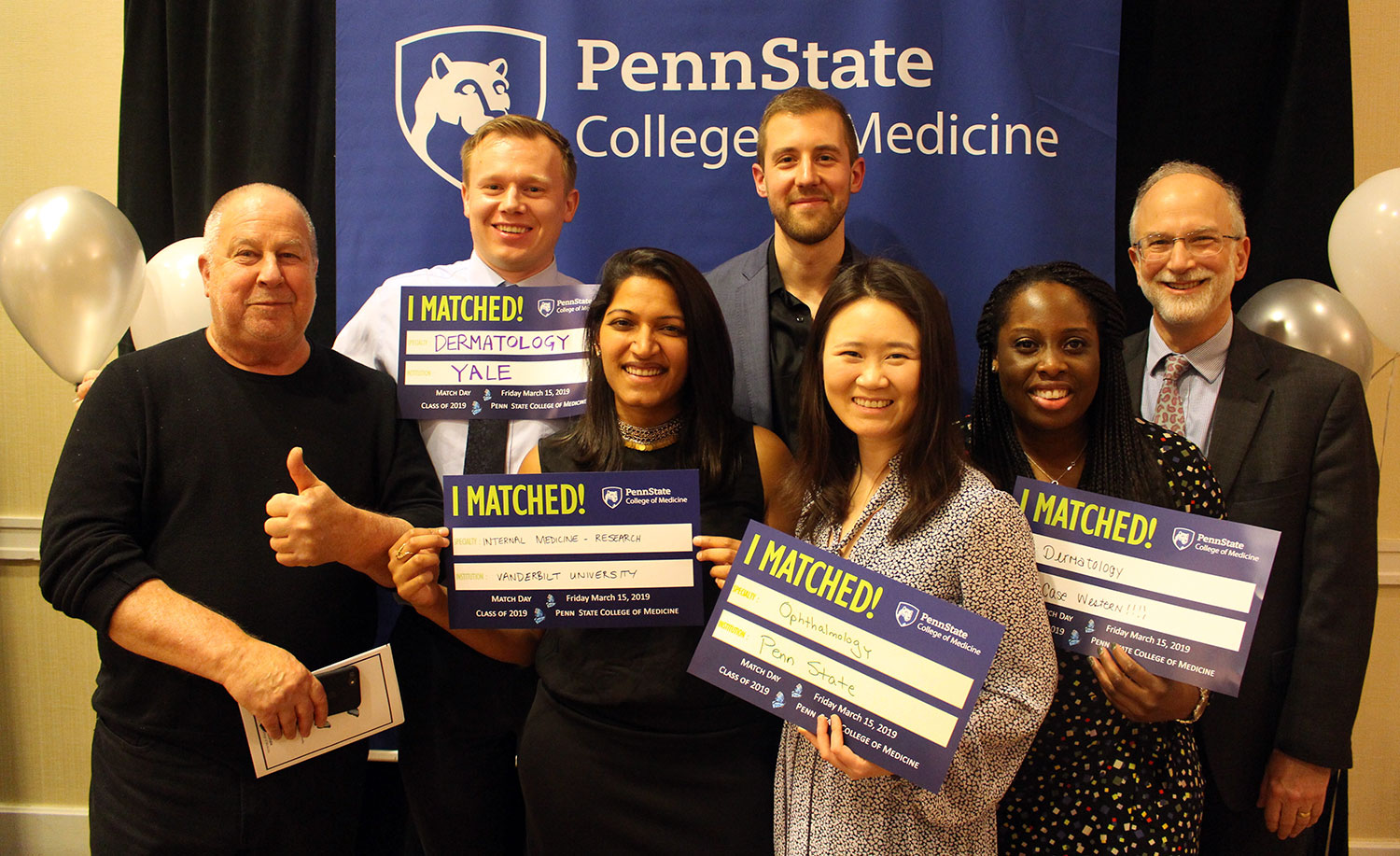 A group of seven people is seen standing in front of a Penn State College of Medicine banner. The students in the middle are holding signs that say 